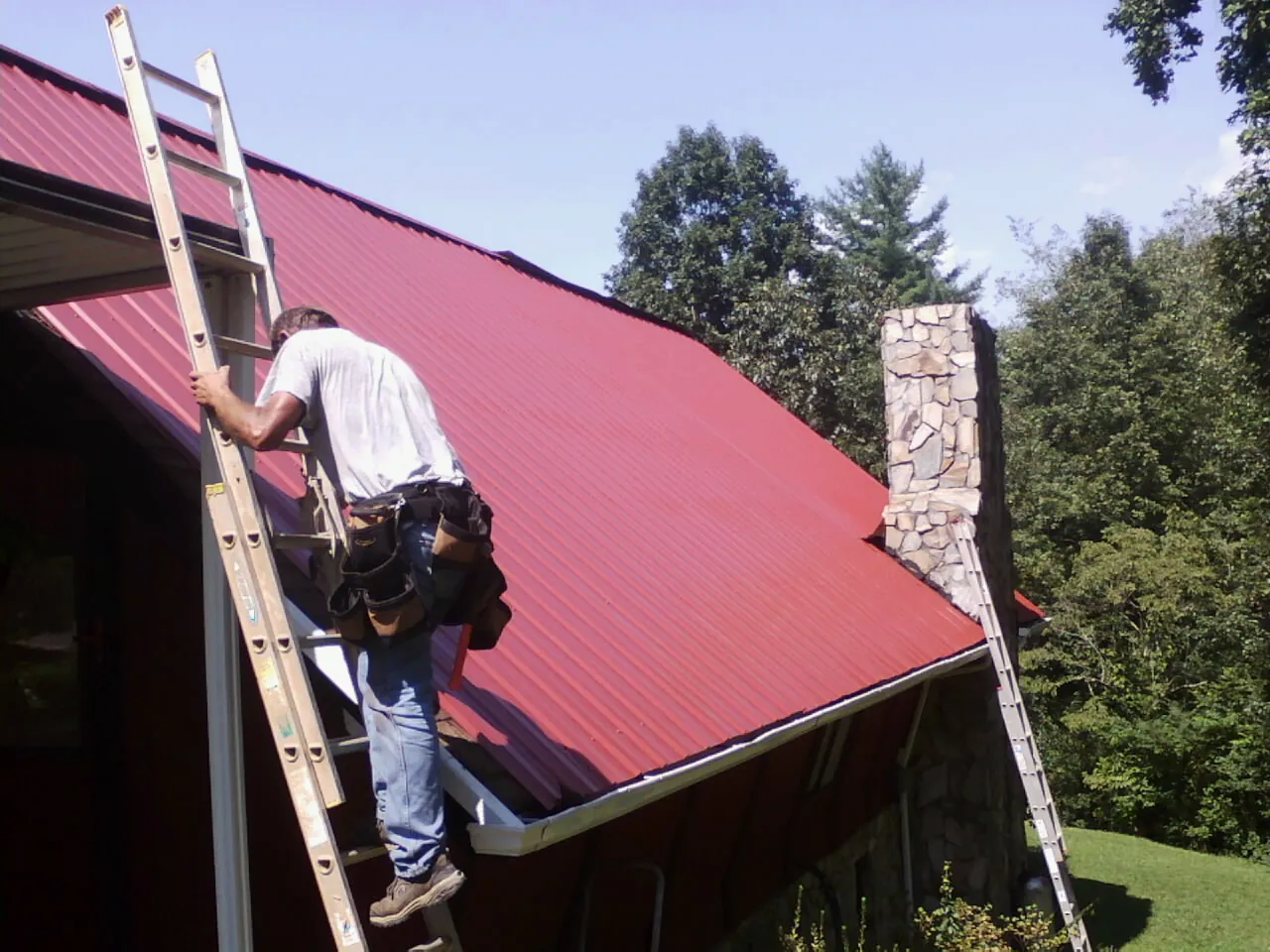 Roofer on a ladder beside a red metal roof and stone chimney — Soffit & Fascia Repair by Roofing Brothers in Normalville